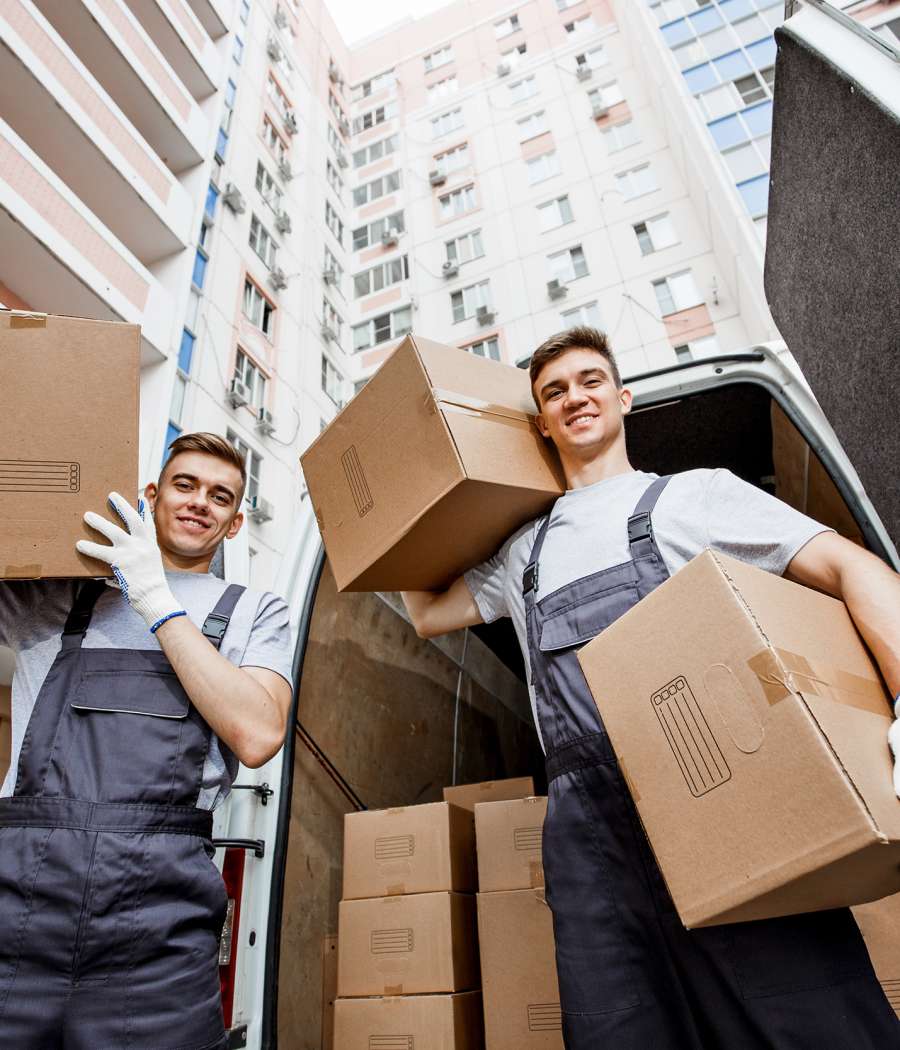 Two young handsome smiling workers wearing uniforms are standing in front of the van full of boxes holding boxes in their hands. The block of flats is in the background. House move, mover service Two young handsome smiling workers wearing uniforms are standing in front of the van full of boxes holding boxes in their hands. The block of flats is in the background. House move, mover service.